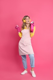Beautiful young woman with headphones, rag and bottle of detergent singing on pink background
