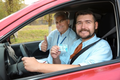Instructor near happy man showing driving license in car