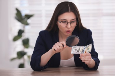 Photo of Expert authenticating 100 dollar banknote with magnifying glass at table in office, focus on hand. Fake money concept