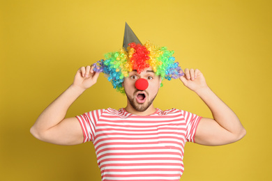 Emotional young man with party hat and clown wig on yellow background. April fool's day