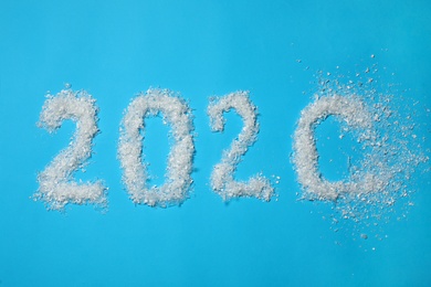Year number 2020 made of artificial snow getting blown away on light blue background, flat lay