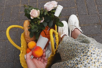 Photo of Woman holding peach near yellow wicker bag of roses, wine and baguette outdoors, above view