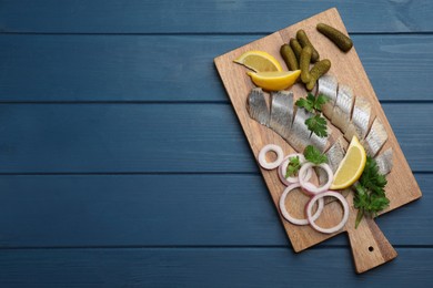 Photo of Sliced salted herring fillet served with parsley, pickles, onion rings and lemon on blue wooden table, top view. Space for text