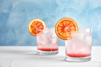 Glasses of drink with ice cubes and orange on table against color background. Space for text