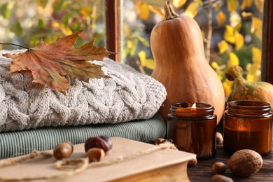 Burning scented candles, warm sweaters, book and pumpkins on wooden table near window. Autumn coziness