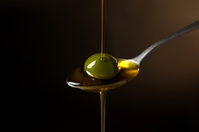 Pouring oil into spoon with ripe olive against dark background, closeup
