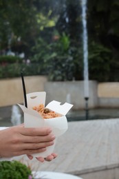 Photo of Woman holding paper box of takeaway noodles with fork outdoors, closeup and space for text. Street food