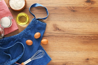 Denim apron with kitchen tool and different ingredients on wooden table, flat lay. Space for text