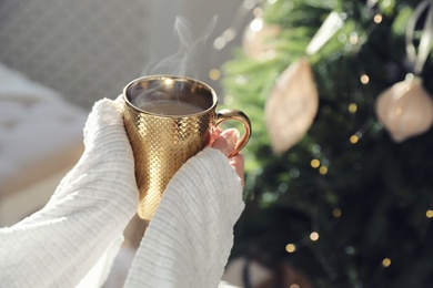 Woman with golden cup of hot drink near Christmas tree at home, closeup