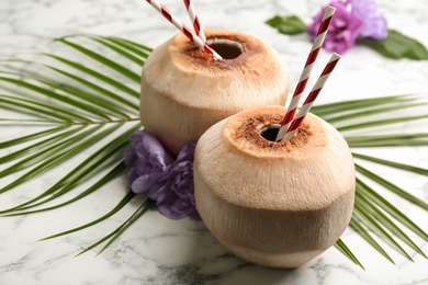 Fresh coconuts with drinking straws and flowers on white marble table, closeup