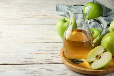 Jug of tasty juice and fresh ripe green apples on white wooden table, space for text