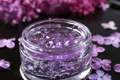 Photo of Jar of cosmetic product and lilac flowers on black surface, closeup