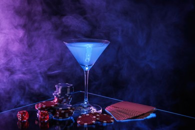 Casino chips, dice, playing cards and cocktail on dark background with smoke, space for text
