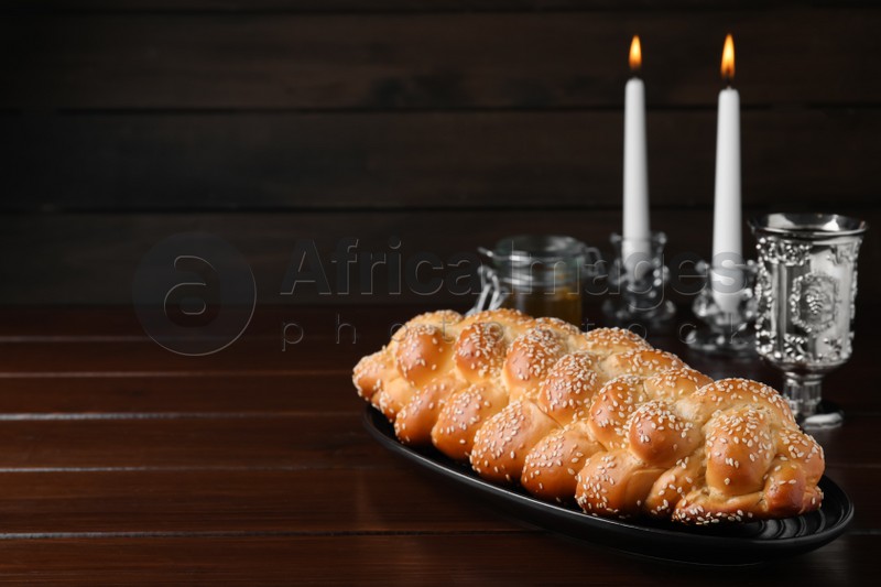 Photo of Homemade braided bread with sesame seeds, goblet and candles on wooden table, space for text. Traditional Shabbat challah