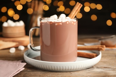 Glass cup of hot cocoa with aromatic cinnamon and marshmallows on wooden table against blurred lights