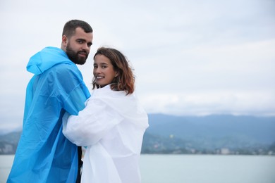 Young couple in raincoats enjoying time together under rain on beach, space for text