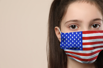 Little girl wearing mask with USA flag pattern on beige background, closeup. Dangerous virus