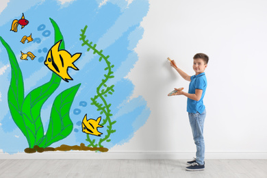 Cute child drawing sea with tropical fishes on white wall indoors