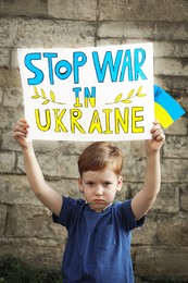 Sad boy holding poster Stop War In Ukraine and national flag against brick wall outdoors