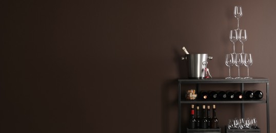 Rack with bottles of wine and glasses near brown wall, space for text