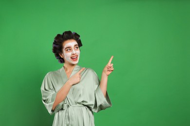 Happy young woman in silk bathrobe with hair curlers and facial mask on green background, space for text