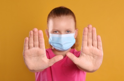 Little girl in protective mask showing stop gesture on yellow background. Prevent spreading of coronavirus