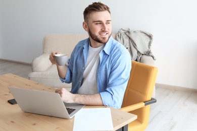 Photo of Young man drinking coffee while working with laptop at desk. Home office