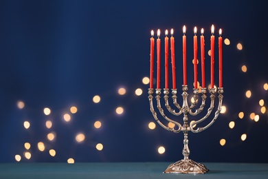 Silver menorah with burning candles on table against blue background and blurred festive lights, space for text. Hanukkah celebration