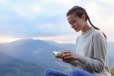 Beautiful young woman reading book in mountains, space for text