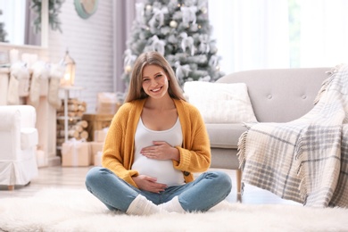 Young pregnant woman sitting on floor in room decorated for Christmas
