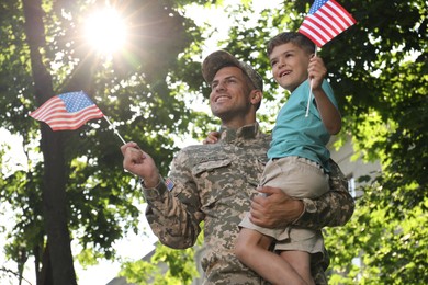 Soldier and his little son with American flags outdoors, low angle view. Veterans Day in USA