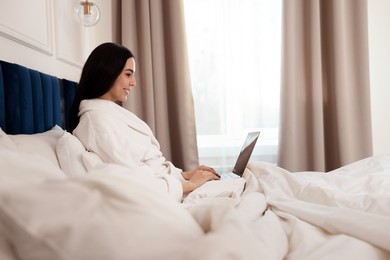 Happy young woman working with laptop on bed in hotel room