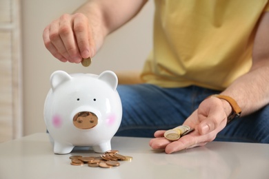 Man putting coin into piggy bank at table indoors, closeup. Money saving