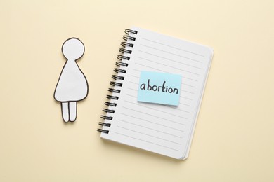 Notebook with word Abortion and pregnant woman paper cutout on beige background, flat lay