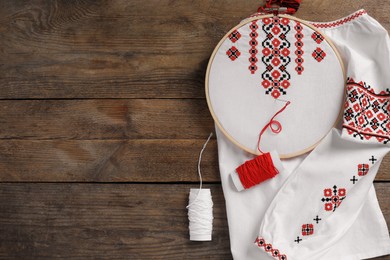 Shirt with red Ukrainian national embroidery in hoop, needle and threads on wooden table, flat lay. Space for text