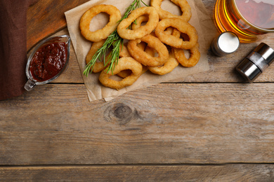 Delicious crunchy fried onion rings, sauce and beer on wooden table, flat lay. Space for text