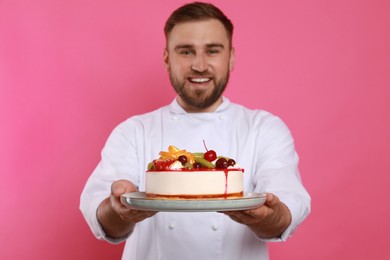 Happy professional confectioner in uniform holding delicious cake on pink background