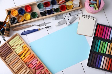 Blank sheet of paper, colorful chalk pastels and other drawing tools on white wooden table, flat lay. Modern artist's workplace