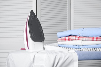 Photo of Modern iron, clean shirt and folded clothes on board indoors