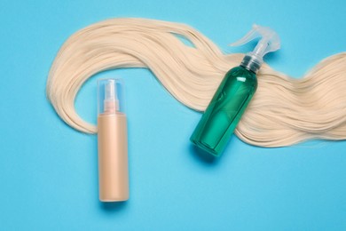 Photo of Spray bottles with thermal protection and lock of blonde hair on light blue background, flat lay