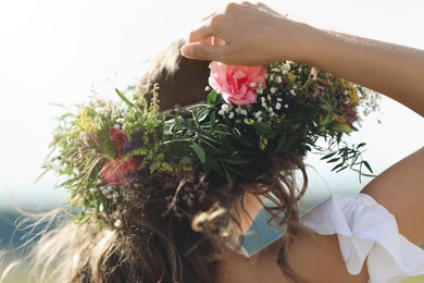 Photo of Young woman wearing wreath made of beautiful flowers outdoors on sunny day, closeup