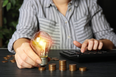 Woman with light bulb, calculator and coins at black wooden table, closeup. Power saving