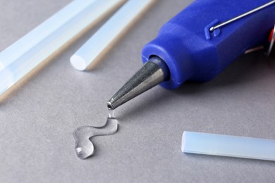 Photo of Melted glue dripping out of hot gun nozzle near sticks on grey background, closeup