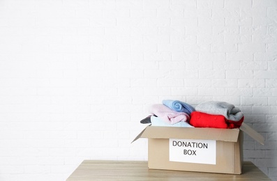 Donation box with knitted clothes on table near brick wall. Space for text