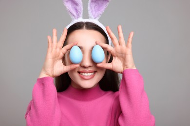 Photo of Happy woman in bunny ears headband holding painted Easter eggs near her eyes on grey background