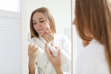 Young woman with acne problem holding bottle with cosmetic product in bathroom