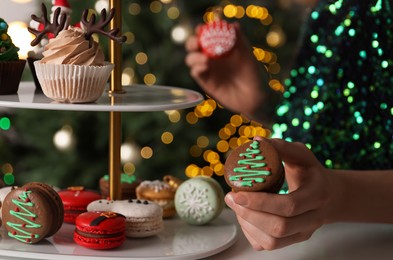 Woman taking Christmas macaron from stand against blurred festive lights, closeup