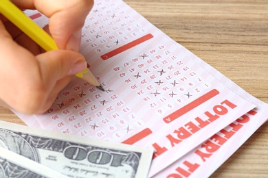 Woman filling out lottery tickets with pencil and money on wooden table, closeup. Space for text