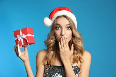 Surprised young woman wearing Santa hat with Christmas gift on light blue background