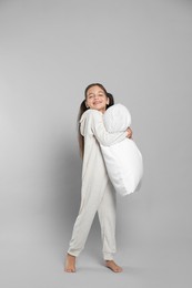 Cute girl in white pajamas hugging pillow on light grey background